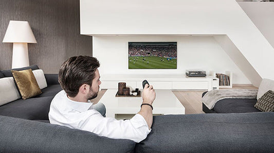 Een meneer op een bank die de televisie bedient met een afstandsbediening terwijl de apperatuur is geplaatst in het TV meubel